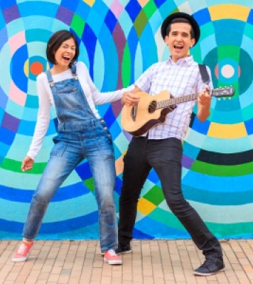 A woman in overalls and a man with a hat playing a guitar standing by each other in front of a colorful wall