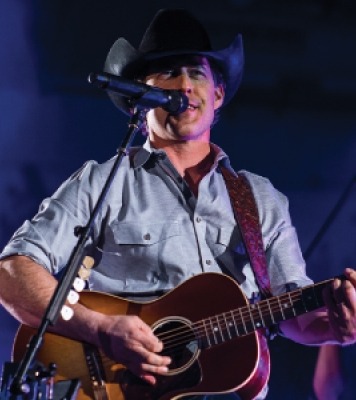 Man with a button down shirt and a cowboy hat playing a guitar and singing into a microphone