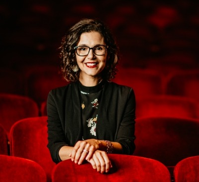 Woman with brown hair, glasses, black blazer sitting in red theater seats