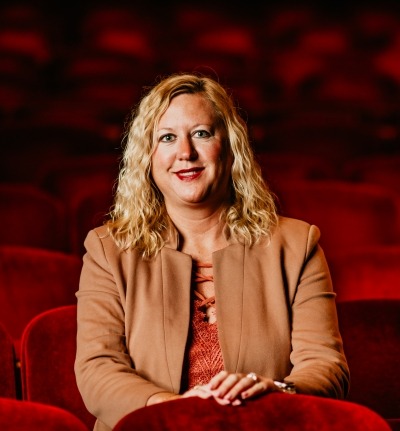 Woman with loose curls blonde hair in a tan leather jacket sitting in red theater seats